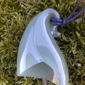 May include: A light green jade pendant carved in the shape of a wave, hanging from a black cord. The pendant is smooth and polished, with a subtle sheen.