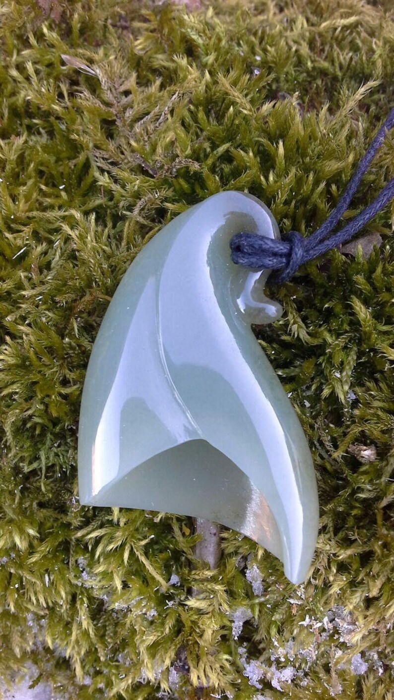 May include: A light green jade pendant carved in the shape of a wave, hanging from a black cord. The pendant is smooth and polished, with a subtle sheen.
