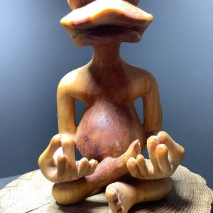 May include: A wooden frog sculpture sitting in a meditative pose with its eyes closed. The frog is carved from a light brown wood with a reddish hue. The frog is sitting on a wooden stump.