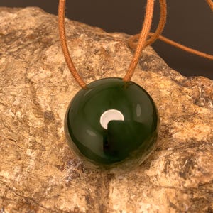 May include: A dark green, glossy, round pendant necklace with a light brown cord. The pendant has a white crescent-shaped design. The necklace is displayed on a textured, light brown stone surface.
