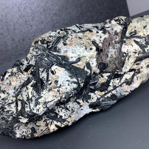 Puede incluir: Espécimen de piedra natural con una mezcla de colores negro, blanco y gris. La piedra tiene una superficie rugosa y texturizada con formaciones cristalinas. Los cristales negros están dispersos por la matriz más clara.