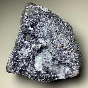 May include: A rough, gray rock with a large, irregular cavity filled with clusters of small, purple crystals. The crystals are surrounded by a white, powdery substance.