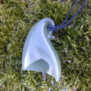 May include: A green jade pendant carved in the shape of a wave, hanging from a black cord. The pendant is smooth and polished, with a subtle sheen.