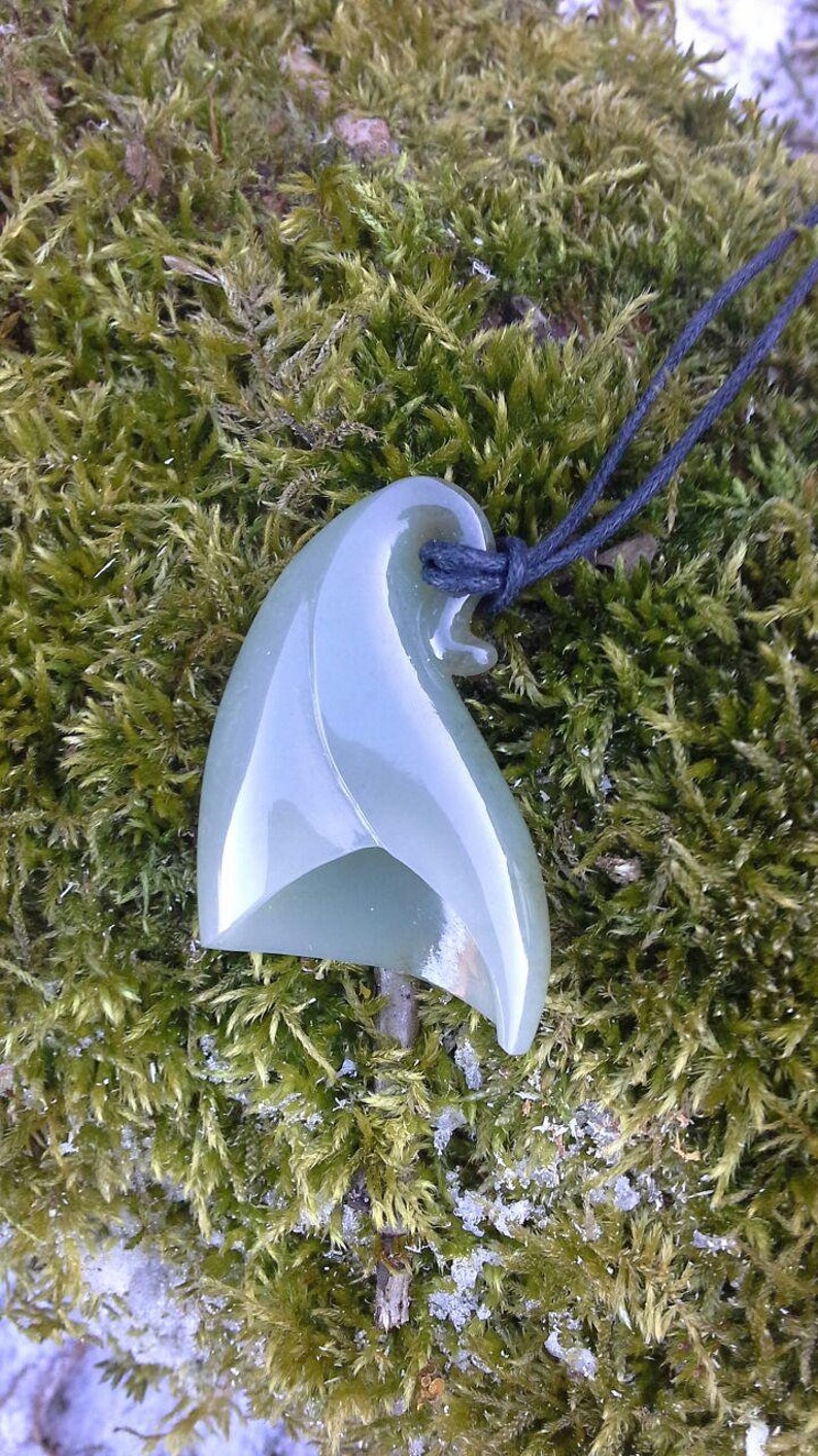 May include: A green jade pendant carved in the shape of a wave, hanging from a black cord. The pendant is smooth and polished, with a subtle sheen.