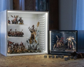 May include: Two white display cabinets with glass doors and shelves filled with miniature painted figures. The figures are arranged in rows and depict various characters and creatures. The cabinets are lit from within, highlighting the details of the figures. There are also several dice scattered on the table in front of the cabinets.