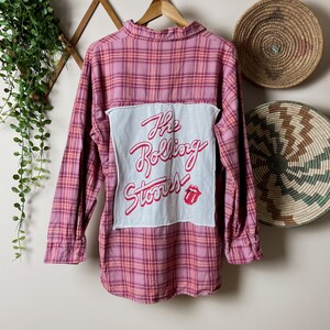 May include: A pink and orange plaid flannel shirt with a white patch sewn on the back. The patch has the words "The Rolling Stones" written in red cursive with a red tongue and lips logo.