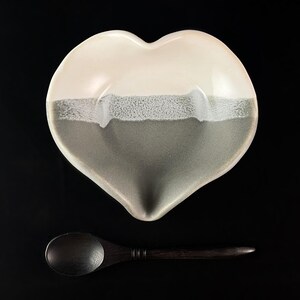 May include: A heart-shaped ceramic dish with a white top and a gray bottom. The dish has a textured glaze and comes with a wooden spoon.