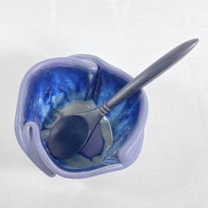 May include: A small, hand-painted ceramic bowl with a blue and purple glaze. The bowl is shaped like a hand holding a wooden spoon. The spoon is also painted blue.