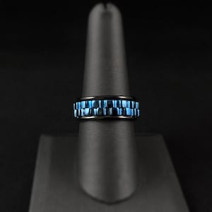 May include: A black ring with a blue, checkered design. The ring is displayed on a black stand against a dark background. The blue color has a metallic sheen.