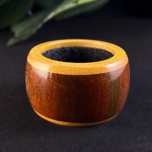 May include: A handcrafted wooden ring with a dark brown band and a light brown rim. The interior is lined with a dark gray material. The ring is a unique piece of jewelry.