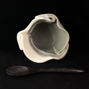 May include: A white and green ceramic bowl with a unique folded design. The bowl is accompanied by a wooden spoon.