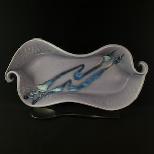 May include: A lavender ceramic serving dish with a blue and white glaze design. The dish has a wavy, organic shape with a spoon resting beside it.