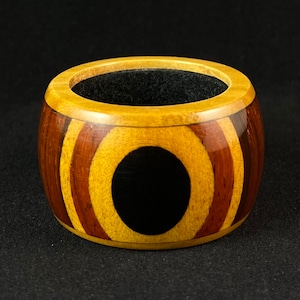 May include: A handcrafted wooden ring box with a circular design. The box features alternating bands of light and dark wood, with a central black circle. The interior is lined with black fabric.