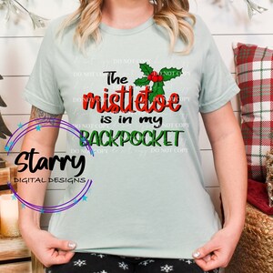 Könnte beinhalten: Hellgrünes T-Shirt mit dem Text "The mistletoe is in my BACKPOCKET" in Rot, Grün und Schwarz. Das Design beinhaltet einen Mistelzweig mit roten Beeren. Ein weihnachtliches Kleidungsstück.