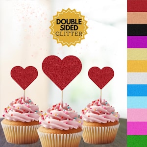 May include: Three cupcakes with pink frosting and colourful sprinkles topped with red glitter heart cupcake toppers. The toppers are double-sided glitter and say "Double Sided Glitter" on the label.