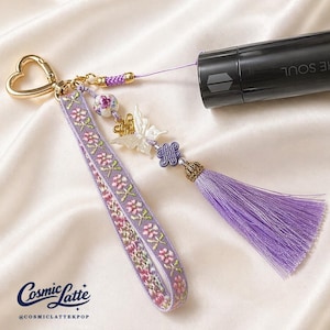 Könnte beinhalten: Ein lila-goldener Schlüsselanhänger mit herzförmigem Verschluss und einem floral bestickten Handgelenkband. Der Schlüsselanhänger hat eine Quaste, einen Schmetterlingsanhänger und eine dekorative Perle. Der Text "THE SOUL" ist auf einem schwarzen Objekt sichtbar.