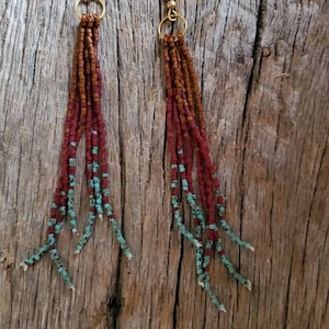 May include: A pair of long, beaded earrings with a gradient of brown and turquoise beads. The earrings are hanging from gold hooks.