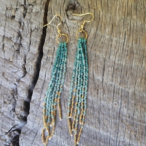 May include: Beaded earrings with a fringe design. The earrings have teal and gold beads, with gold-coloured ear wires. The beads are arranged vertically in a fringed pattern, creating a flowing, textured look. The earrings are displayed against a wooden background.