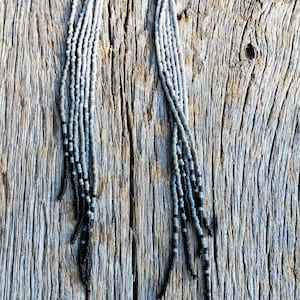May include: A pair of long, beaded earrings with a gradient of gray and black beads. The earrings are hanging on a piece of weathered wood.