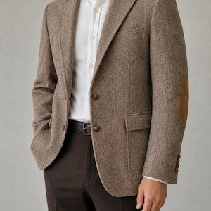 May include: A textured brown blazer featuring a white button-down shirt underneath, dark brown trousers, and a black belt. The blazer has two front buttons, two flap pockets, and brown suede elbow patches on the sleeves with small brown buttons.