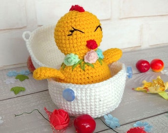 May include: A yellow crocheted chick with a red beak and closed eyes, wearing a flower crown, sits inside a white crocheted egg.
