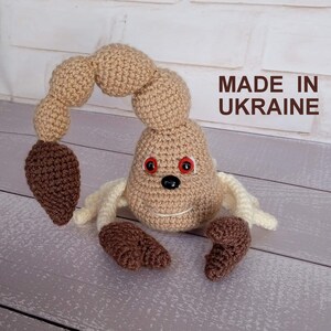 May include: A handmade crochet scorpion toy in tan, brown, and cream colors. The scorpion has a curved tail, red eyes, and a smiling face. The text "MADE IN UKRAINE" is visible.