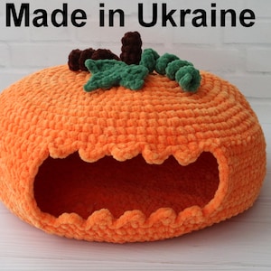Pode incluir: Uma cama para animais de estimação laranja, feita de crochê em forma de abóbora. A cama tem uma abertura ondulada e é adornada com folhas verdes e caules marrons. O texto "Made in Ukraine" está no topo.