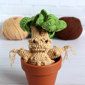 May include: A crocheted plush toy of a mandrake plant in a terracotta pot. The plant has a green leafy top and a beige body with a face. The toy is sitting on a white wooden surface.