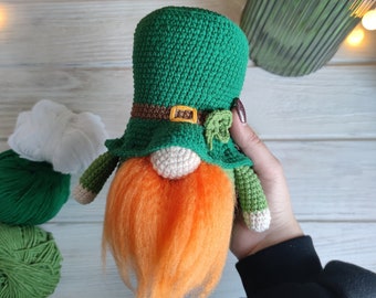May include: A green crocheted gnome with an orange beard and a green hat with a brown belt. The gnome is being held by a hand.