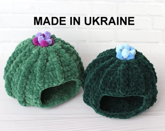 May include: Two green crocheted pet beds shaped like a cactus with a flower on top. One bed has a purple flower and the other has a blue flower. The beds are made in Ukraine.