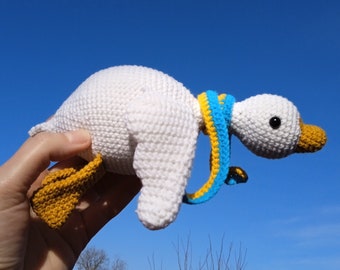 May include: A white crocheted goose toy with yellow and blue crocheted accents. The goose is wearing a blue and yellow scarf around its neck. The goose is being held up in the air by a hand.