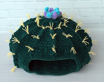 May include: A green crocheted cactus pet bed with yellow yarn spikes and a purple and blue flower on top.