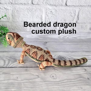 Peut inclure: Un jouet en peluche en crochet en forme de dragon barbu, avec un schéma de couleurs brun clair, beige et vert, est posé sur une surface en bois. Le jouet est une peluche personnalisée et est fait pour ressembler à un vrai dragon barbu.