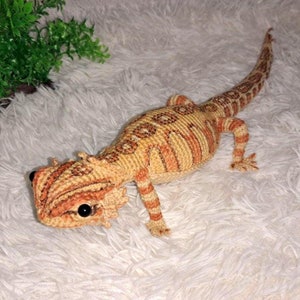 以下が含まれることがあります： オレンジと茶色のストライプのついた、かぎ針編みで作られたヒゲトカゲのおもちゃ。おもちゃは黒い目と質感のある表面をしています。