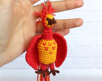 May include: A crocheted red and yellow phoenix keychain with a brown beak and legs. The phoenix has a long tail with red and yellow yarn strands.