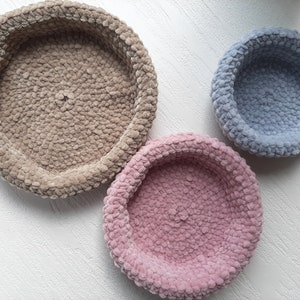 May include: Three round, crocheted,  cushioned bowls in different sizes. The largest bowl is beige, the medium bowl is pink, and the smallest bowl is light blue.