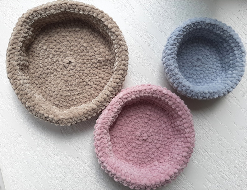 May include: Three round, crocheted,  cushioned bowls in different sizes. The largest bowl is beige, the medium bowl is pink, and the smallest bowl is light blue.