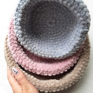 May include: Three crocheted bowls, one light blue, one pink, and one beige, are stacked on top of each other.