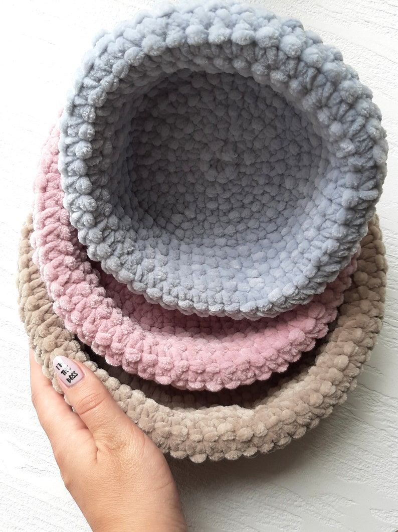 May include: Three crocheted bowls, one light blue, one pink, and one beige, are stacked on top of each other.