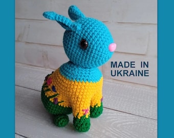 May include: A crocheted blue, yellow, and green bunny rabbit toy with black eyes and a pink nose. The toy is sitting on a white wooden surface. The text "MADE IN UKRAINE" is visible in the background.