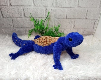 May include: Blue and brown crocheted lizard plush toy with black eyes. The toy is lying on a white fluffy surface.