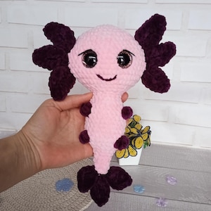 May include: A handmade plush axolotl toy in pink and dark purple. The axolotl has large, sparkly eyes and a smiling face. The plush toy features detailed gills and a tail, and is made of soft, textured material.