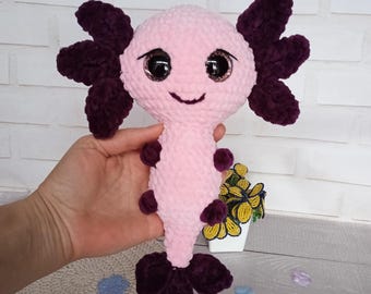 May include: A handmade plush axolotl toy in pink and dark purple. The axolotl has large, sparkly eyes and a smiling face. The plush toy features detailed gills and a tail, and is made of soft, textured material.