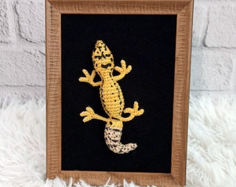 May include: A crocheted leopard gecko figurine in a wooden frame. The gecko is yellow with black spots and a white belly. The frame has a black background.