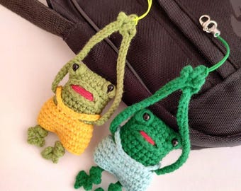 May include: Two crocheted green frog keychains with yellow and blue overalls hanging from a black bag.