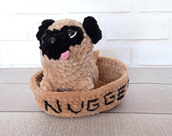 May include: A handmade, plush pug dog toy with black ears and muzzle, a pink tongue, and black eyes, sitting in a brown basket. The basket has the word "NUGGE" embroidered on the side in black thread. The toy is made of soft, textured material.