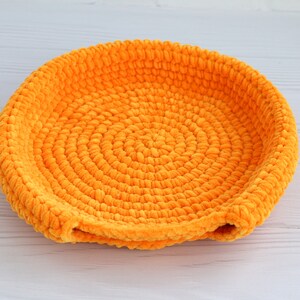 May include: A bright orange crocheted bowl with a circular design. The bowl is made of soft, fuzzy yarn and has a slightly rounded shape.