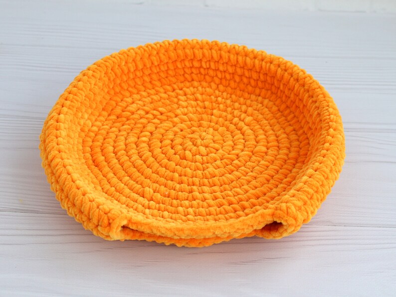 May include: A bright orange crocheted bowl with a circular design. The bowl is made of soft, fuzzy yarn and has a slightly rounded shape.