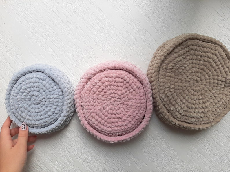 May include: Three round crocheted baskets in different sizes. The largest basket is brown, the middle basket is pink, and the smallest basket is light blue.