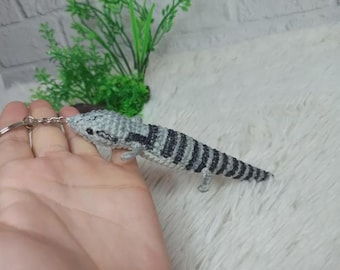 May include: A handmade crochet axolotl keychain. The axolotl is gray with black stripes and small legs. The keychain is attached to a silver metal ring. The background is a white, fluffy surface with green plant accents.